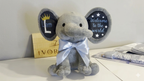 Gray elephant plush toy with a bow tie on a white surface, surrounded by baby items.