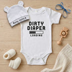 White baby onesie with 'DIRTY DIAPER LOADING' text, white hat, and baby booties on a wooden surface.