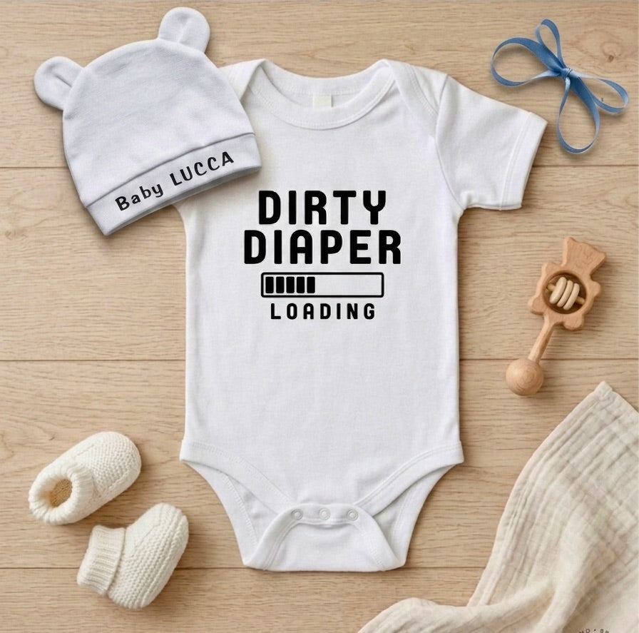 White baby onesie with 'DIRTY DIAPER LOADING' text, white hat, and baby booties on a wooden surface.