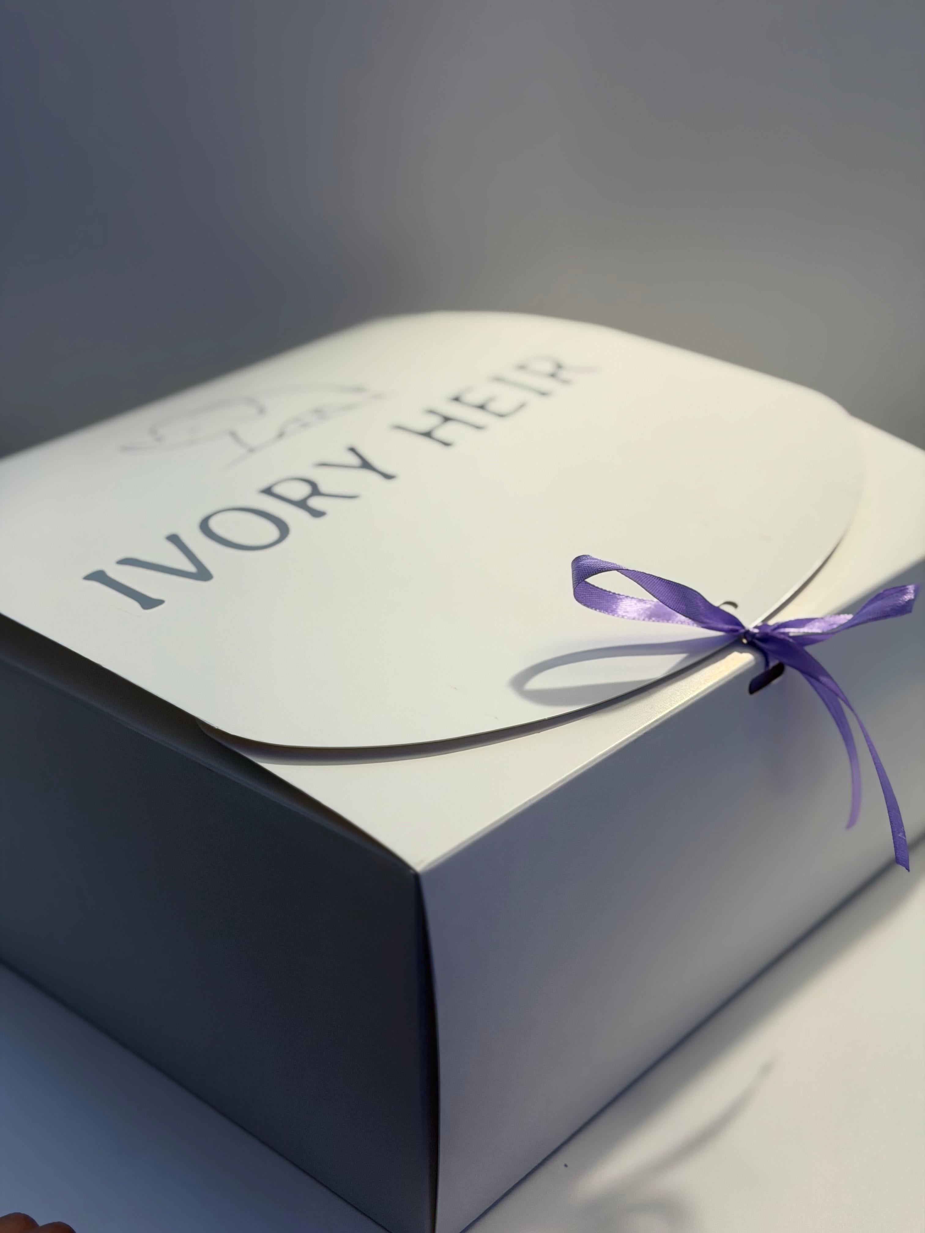 An elegant square gift box in ivory with a matching envelope-style lid printed with the text 'IVORY HEIR' and a small monogram, tied with a purple ribbon bow.