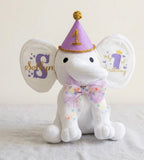 Ivory Heir personalized 12-inch elephant plush toy for a first birthday, featuring a purple party hat and custom stamped monogram ears.