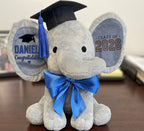 Ivory Heir personalized 9-inch graduation elephant plush toy featuring a black mortarboard cap, royal blue tassel and bow, and custom stamped ears with the name Daniel and Class of 2026.