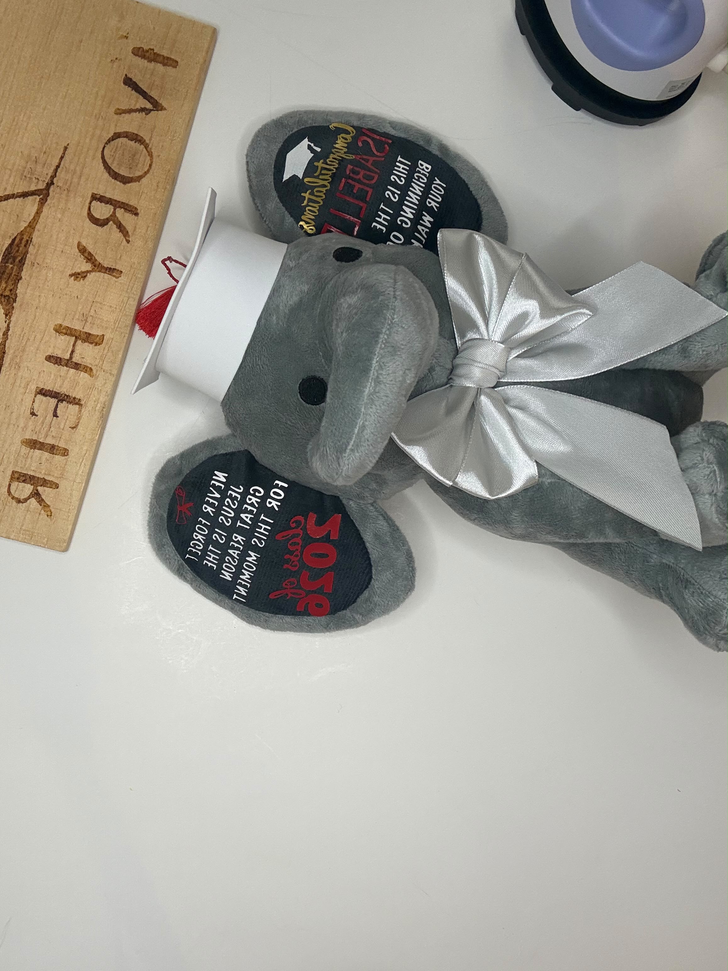 A plush gray elephant toy with a shiny silver bow and a white cup on its trunk