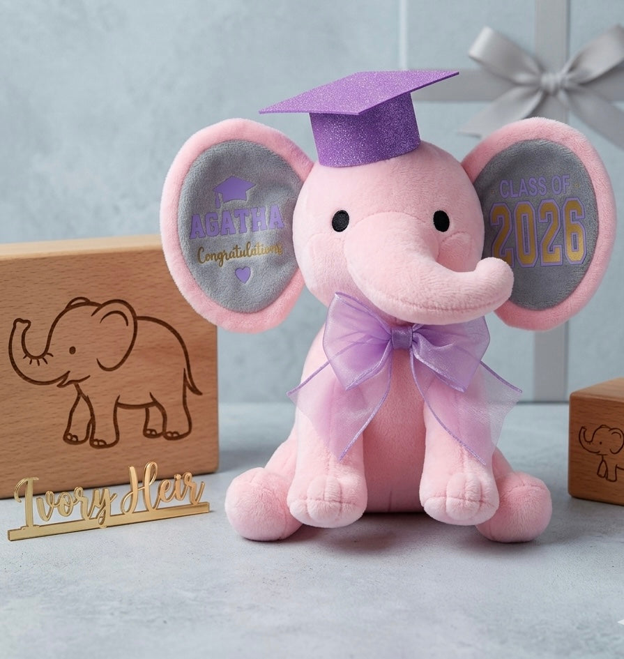 Ivory Heir personalized 9-inch pink graduation elephant plush toy featuring a purple glitter mortarboard cap, matching sheer organza bow, and custom stamped ears with the name Agatha and Class of 2026.