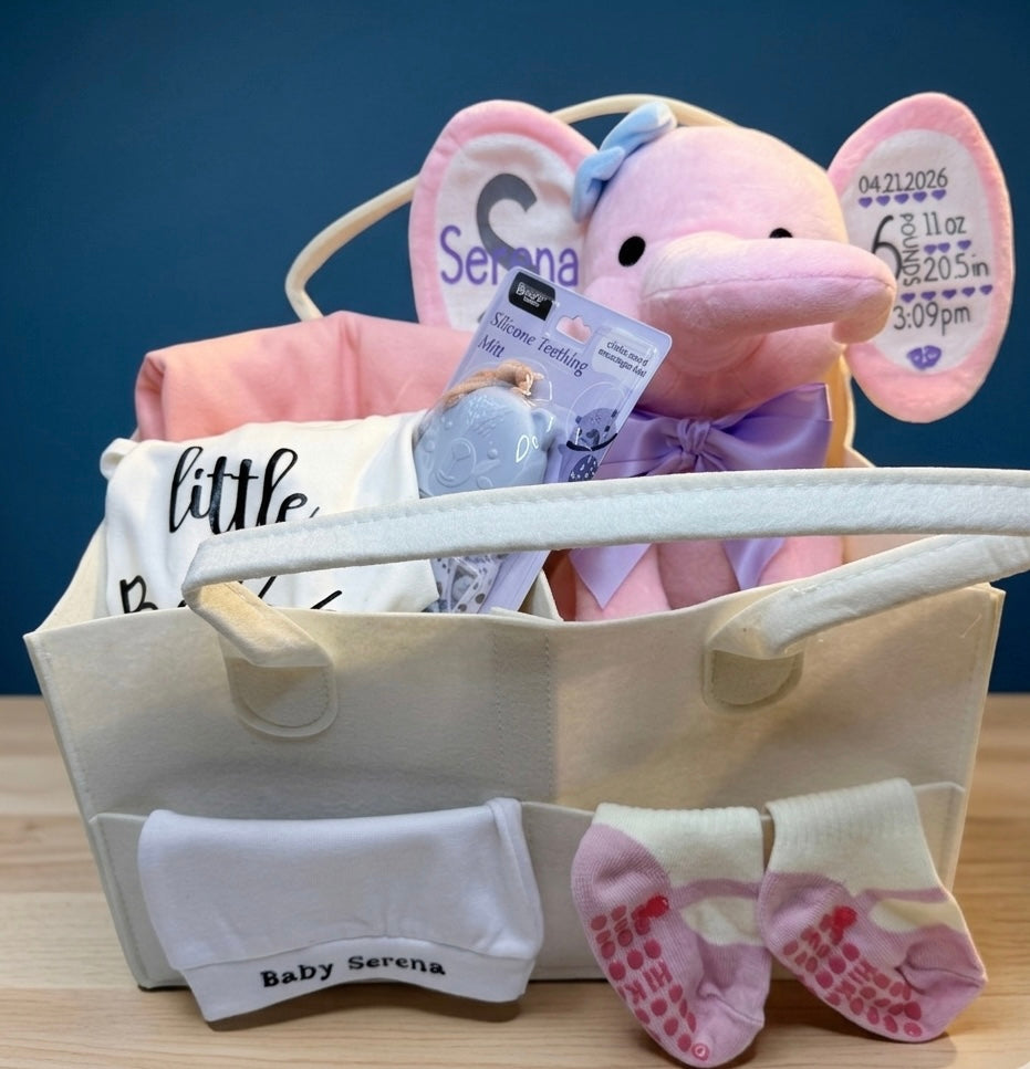 he Grand Welcome Set by Ivory Heir featuring a personalized pink birth stats elephant, custom newborn bodysuit, and luxury baby blanket arranged beautifully in a gift basket.