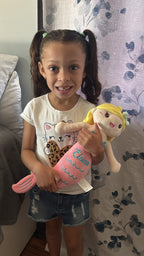 A young girl with pigtails stands indoors holding a pink mermaid plush doll with yellow hair. She is wearing a white graphic t-shirt and denim shorts, standing in front of a patterned curtain.