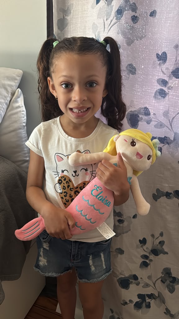 A young girl with pigtails stands indoors holding a pink mermaid plush doll with yellow hair. She is wearing a white graphic t-shirt and denim shorts, standing in front of a patterned curtain.
