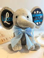 A plush elephant toy with a light gray body, black ears, and a light blue satin bow around the neck. One ear is personalized with the name 'Lucca' in white and blue, and the other reads 'Welcome to the world' with white stars.