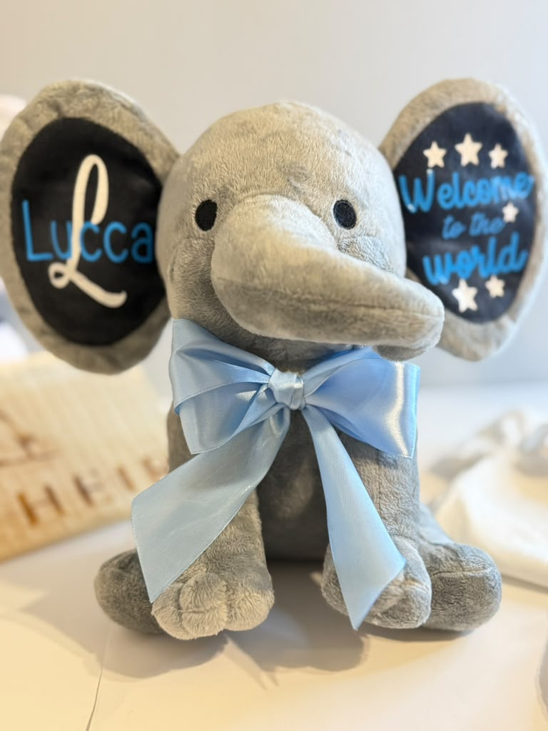 A plush elephant toy with a light gray body, black ears, and a light blue satin bow around the neck. One ear is personalized with the name 'Lucca' in white and blue, and the other reads 'Welcome to the world' with white stars.