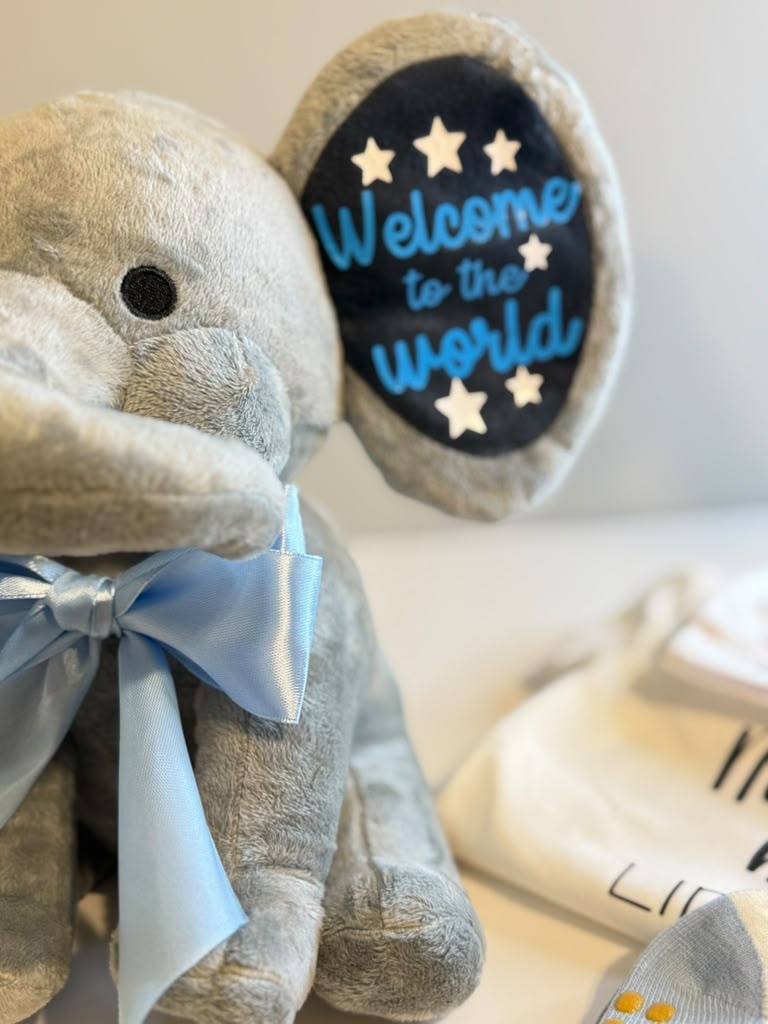 A plush gray elephant toy with a light blue satin bow around its neck and a round black patch on its ear that reads 'Welcome to the world' in blue script with white stars. A folded white garment is partially visible in the background.